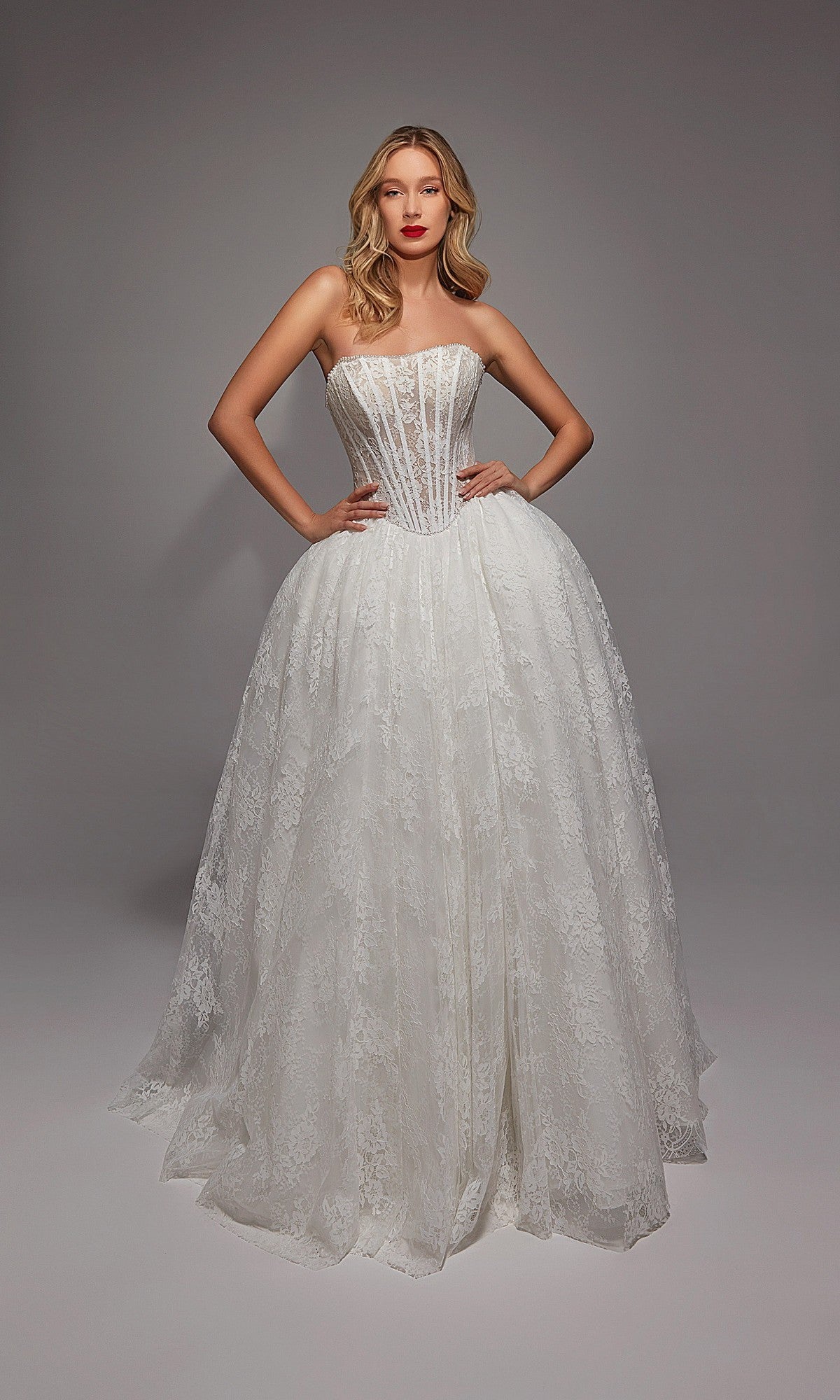 A woman wears the Long White Dress: Alyce 7149, a strapless white gown featuring a full skirt and lace overlay; its fitted bodice showcases classic wedding elegance against a plain gray background.